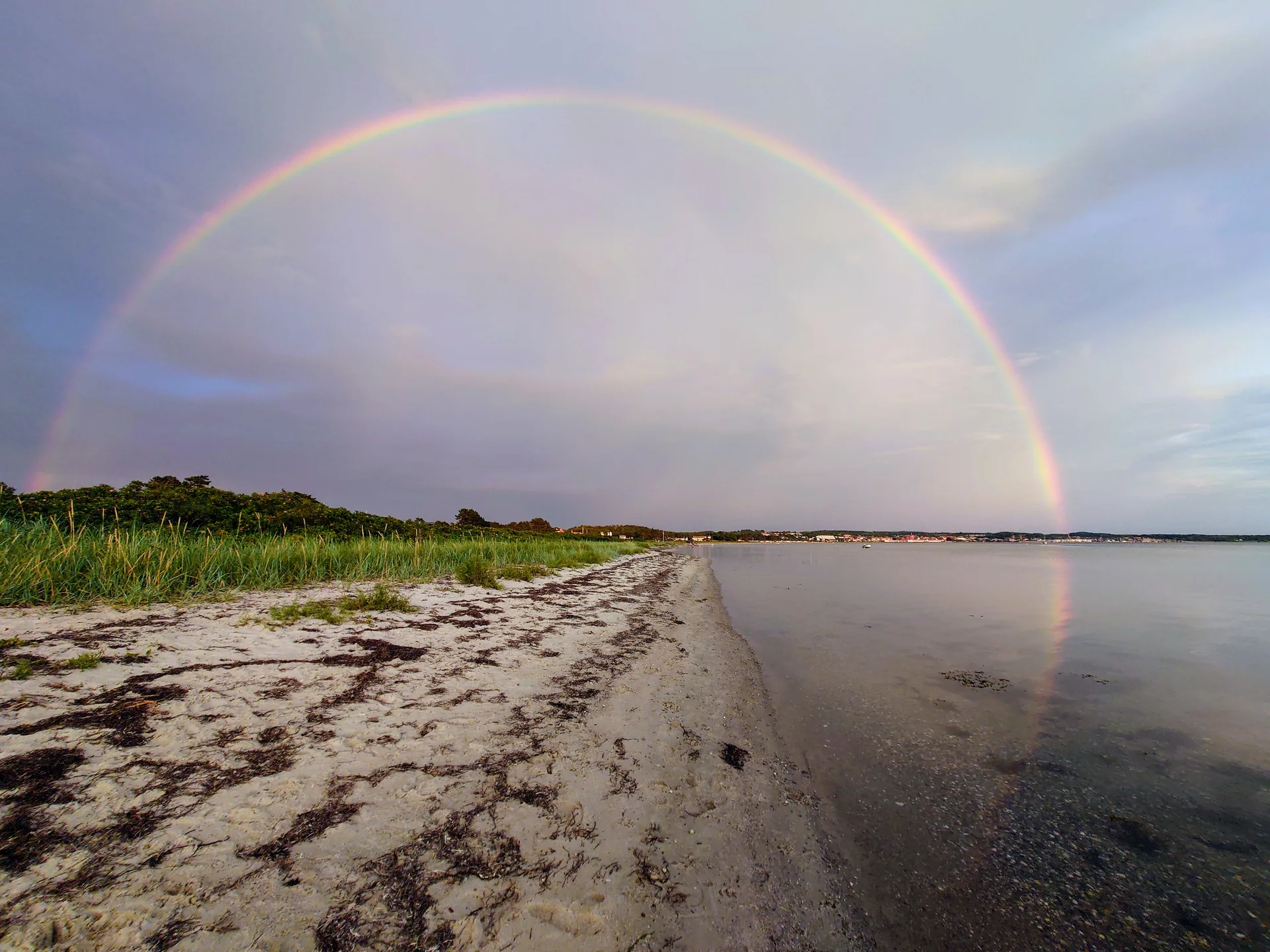 Regnbue over kystlandskab med strand, klitter og roligt vand